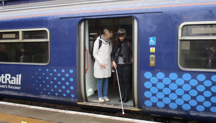 University student with mask covering her eyes attempts to alight from a ScotRail train using a cane with the assistance of a tutor