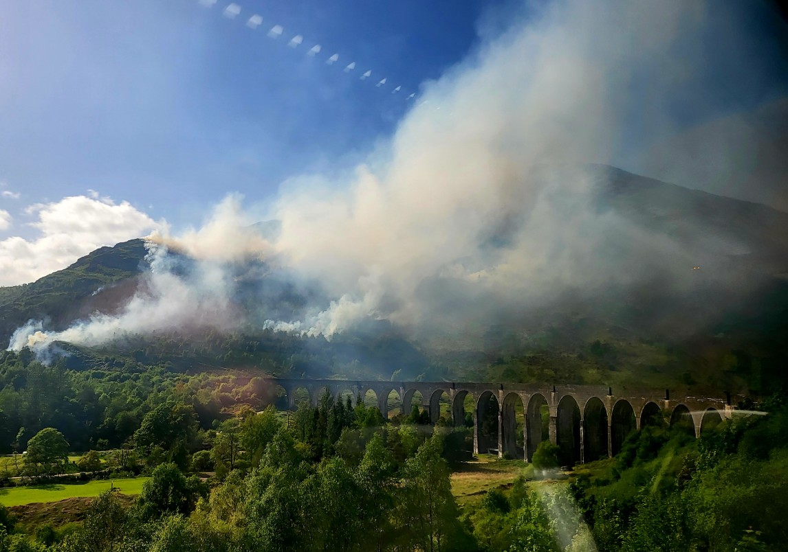 A view out a train window towards the Glenfinnan Viaduct lit up by sunshine while smoke from a wildfire drifts above.