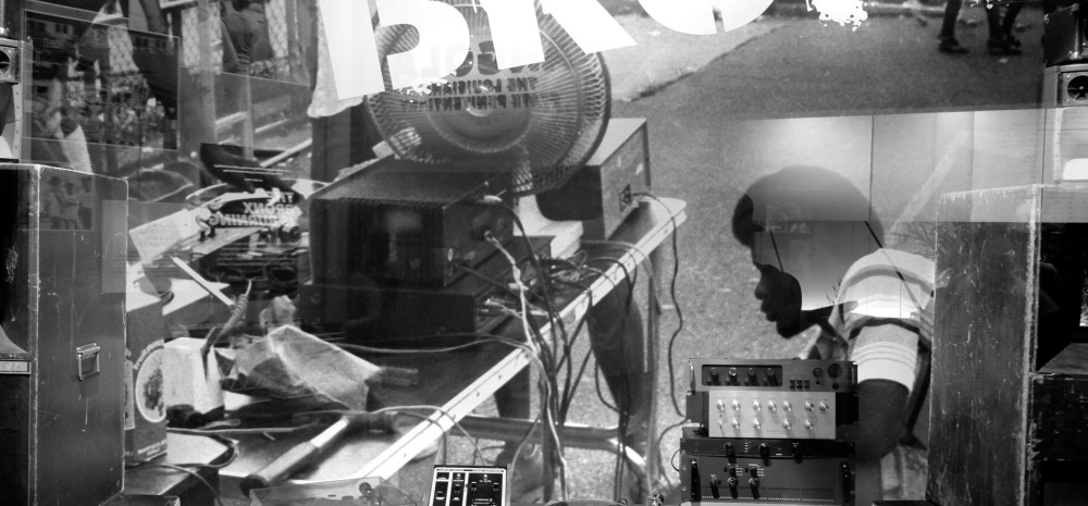 Black and white image of a person sat behind speakers and audio equipment