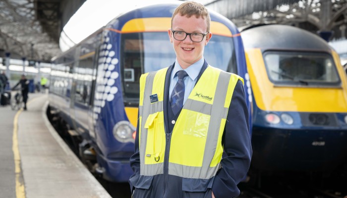 ScotRail Customer Service Modern Apprentice, Ross Henderson, smiling and standing in front of a ScotRail train