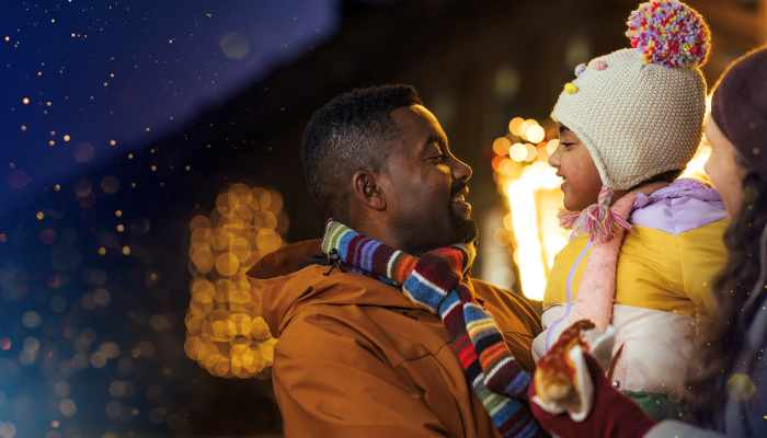 Father lifting and cuddling daughter in an outdoor Christmas setting at night. Both wearing warm clothing and blurred Christmas lights can be seen behind them.